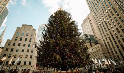 The iconic Rockefeller Christmas Tree against NYC skyscrapers on a sunny winter day.