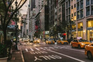 Busy city street of Fifth Avenue lined with tall buildings, yellow taxis, cars and people walking
