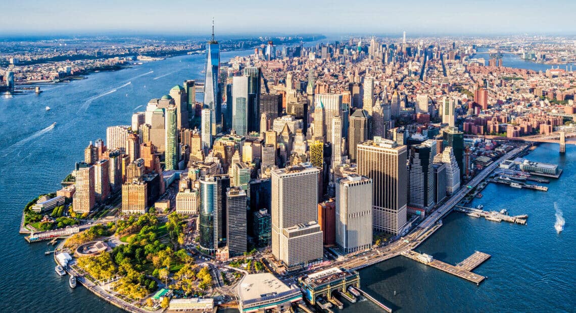 Aerial view of Lower Manhattan at sunset, with the iconic skyscrapers of the Financial District and the World Trade Center lit up against the sky.