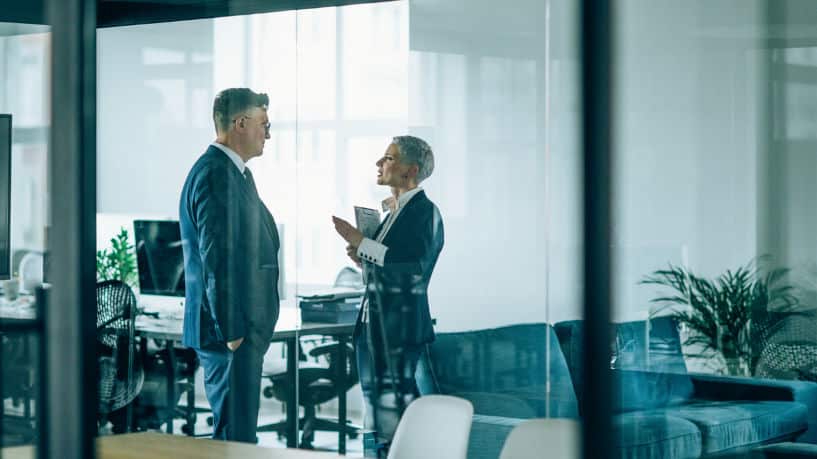 Two lawyers discuss documents in an office, viewed through a glass door