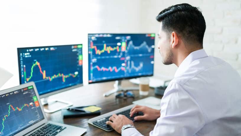 Hispanic male trader typing on a keyboard in office with laptop and dual monitors