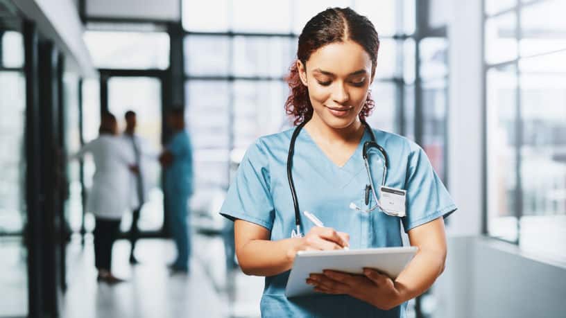 A young nurse is using a digital tablet in a hospital. She is wearing a blue scrubs and has a friendly smile.