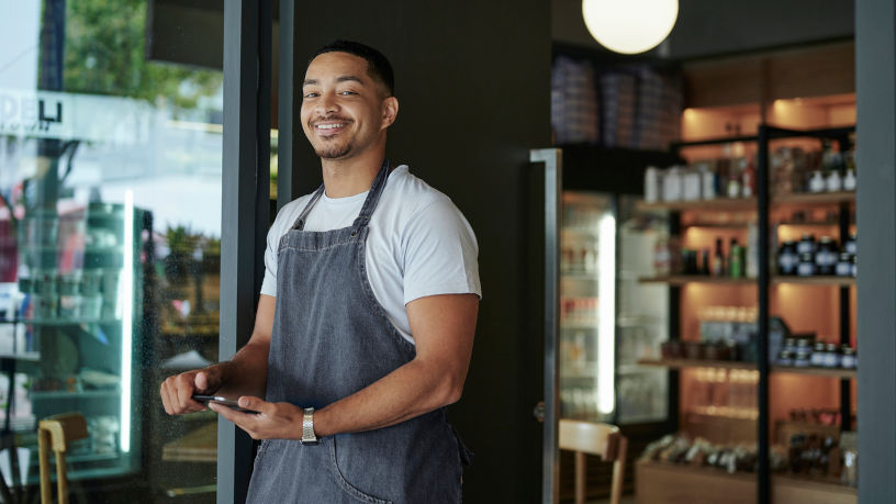 A smiling young male delicatessen owner is standing at the front door of his shop, holding a digital tablet. He is looking at the camera and appears to be happy and confident. The photo is promoting retail space for rent in Manhattan, NYC.