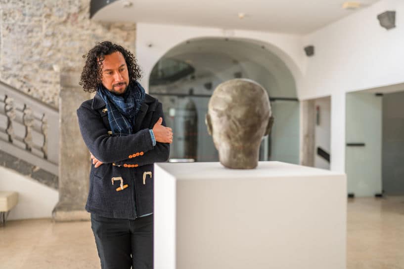 Man in art gallery, black-haired, observing bronze object on pedestal with arms crossed