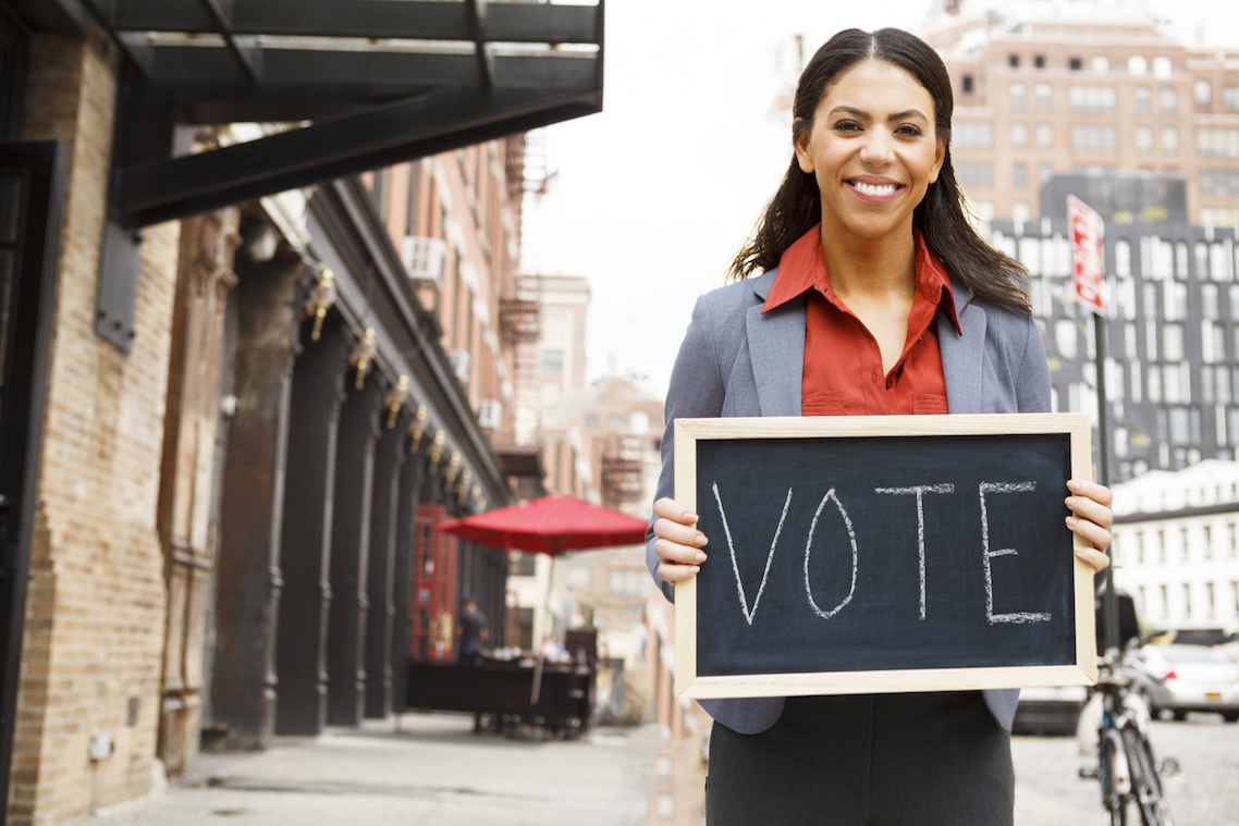 Businesswoman holds "VOTE" sign, smiles on city street.