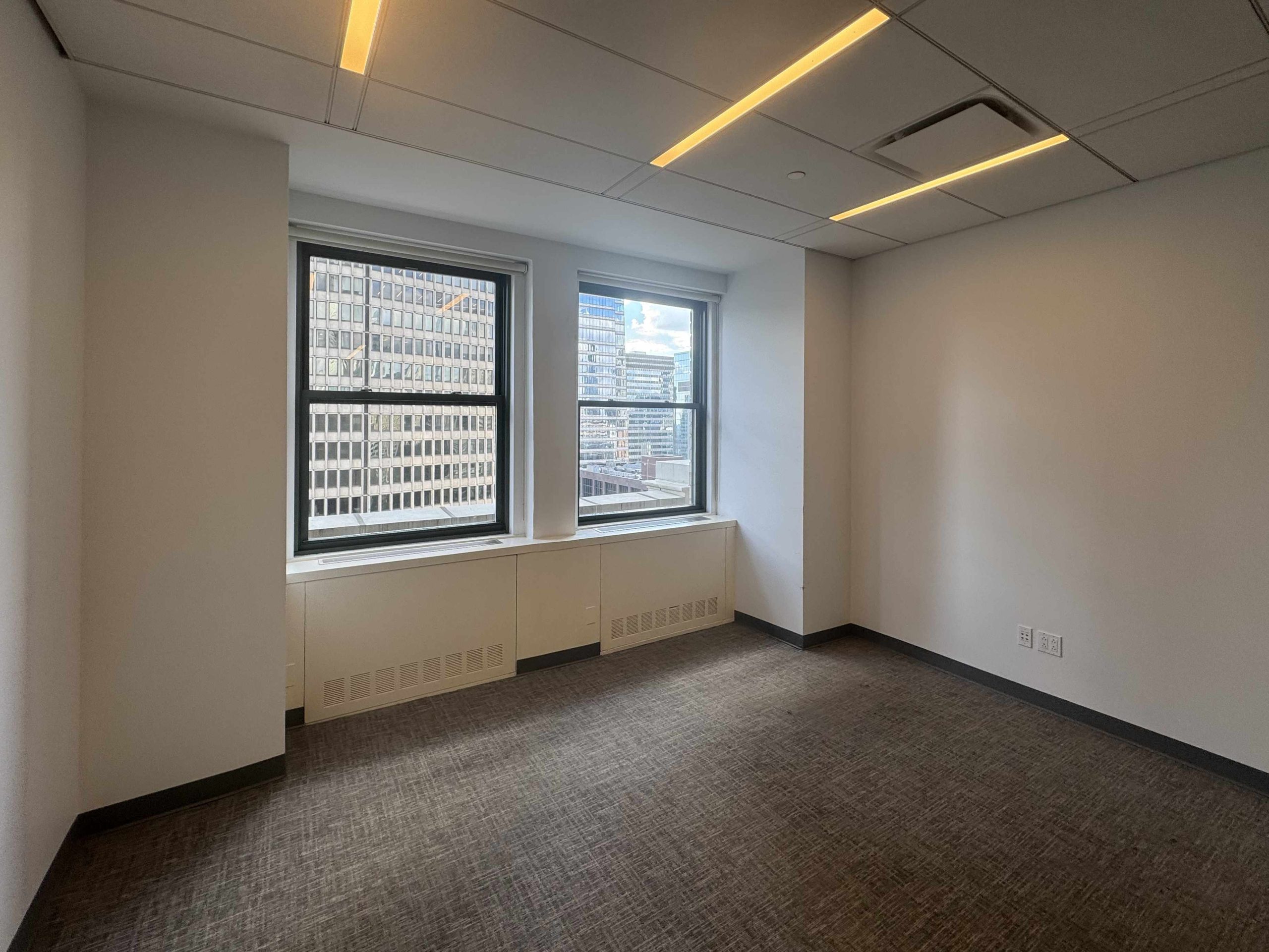 Small office with carpet, big windows, white walls; city skyline visible.