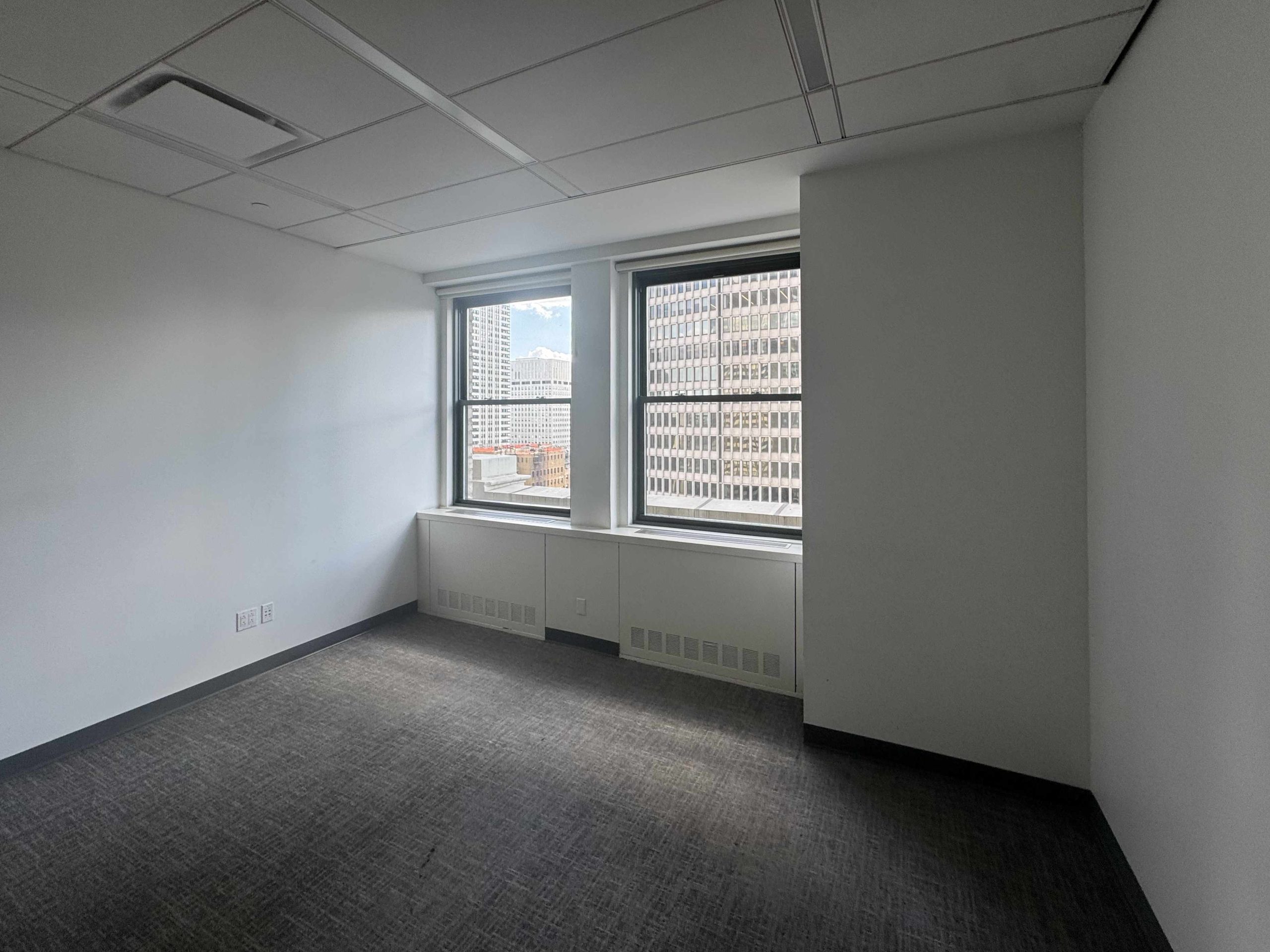 Small empty office with white walls, gray carpet, windows, city view.