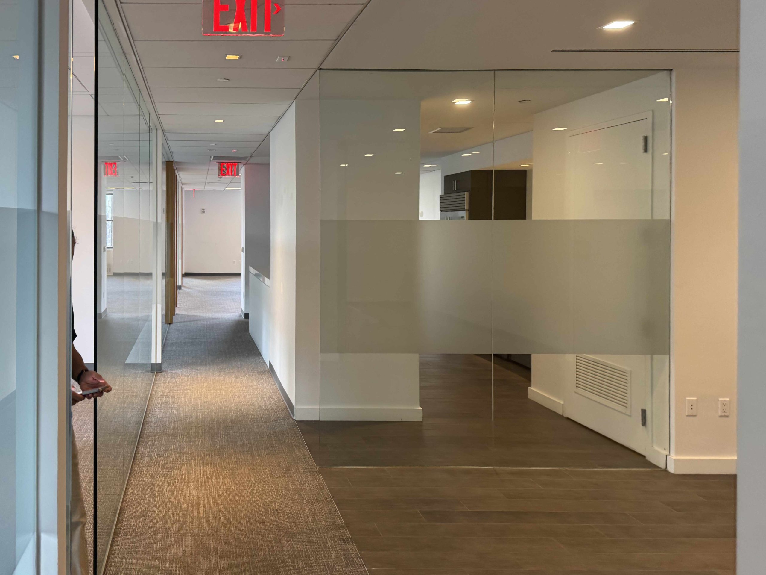 Modern office hallway, glass walls, beige floors, EXIT signs, person on left.