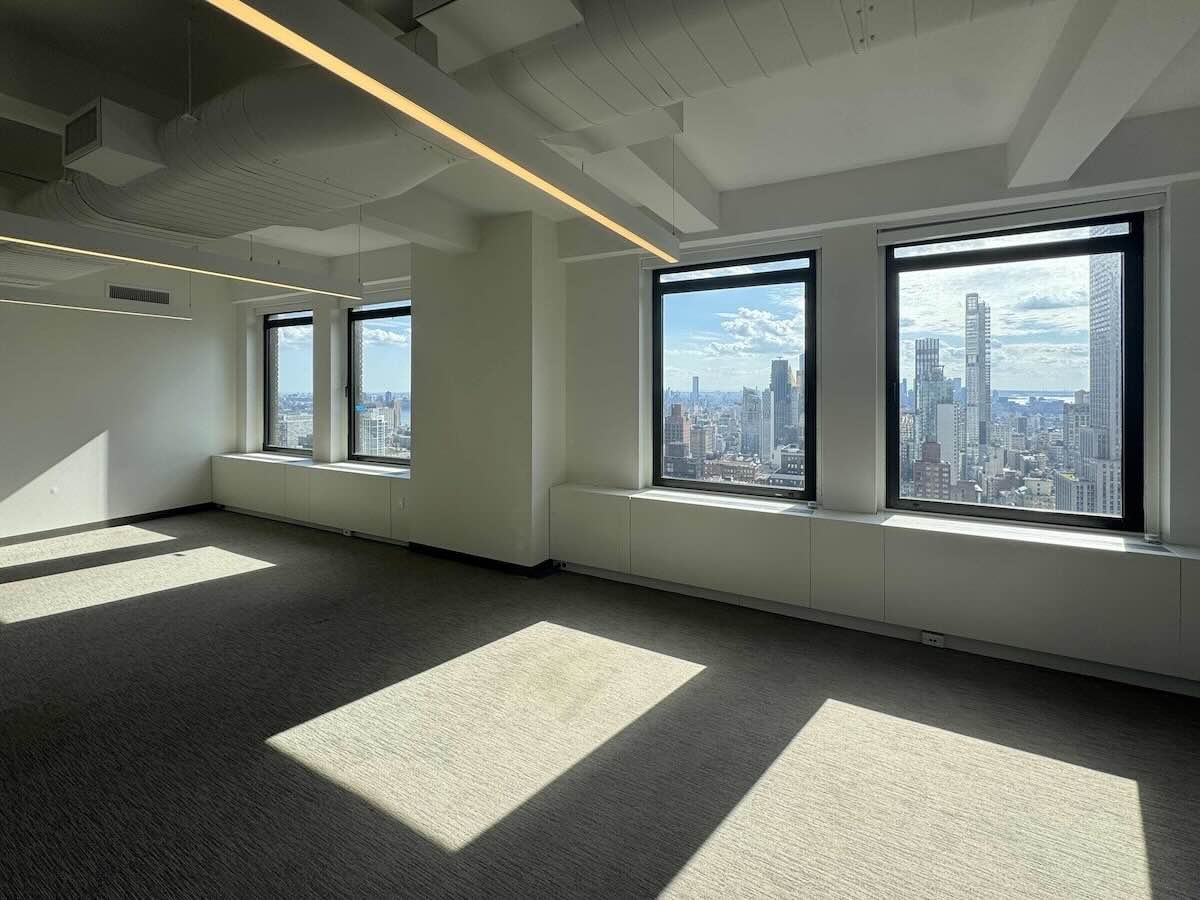 Sunlit empty office with large windows, skyline view, and shadows on carpet.