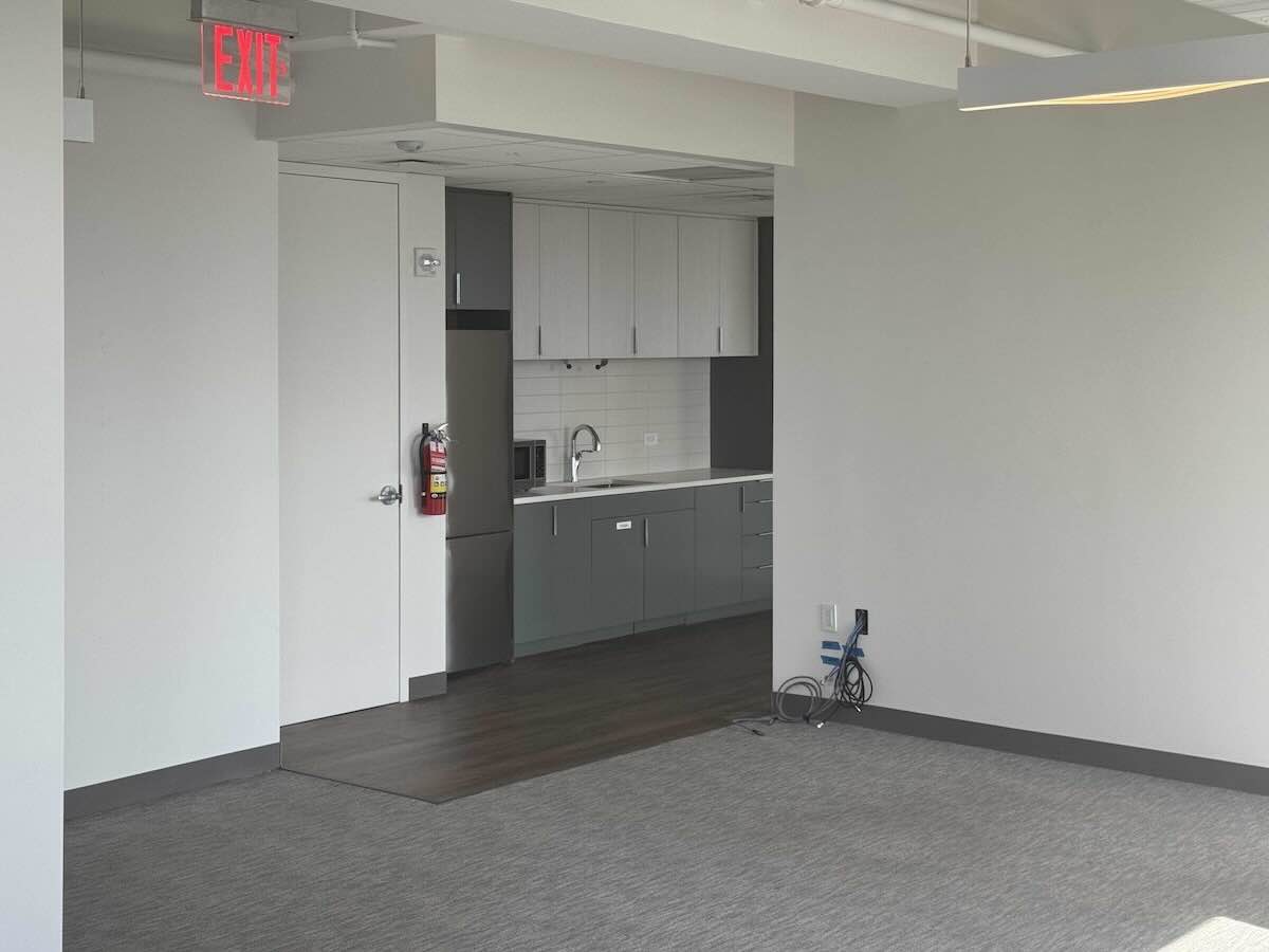 Modern office kitchen, gray cabinets, microwave, fire extinguisher, EXIT sign. Carpeted area with cables in foreground.