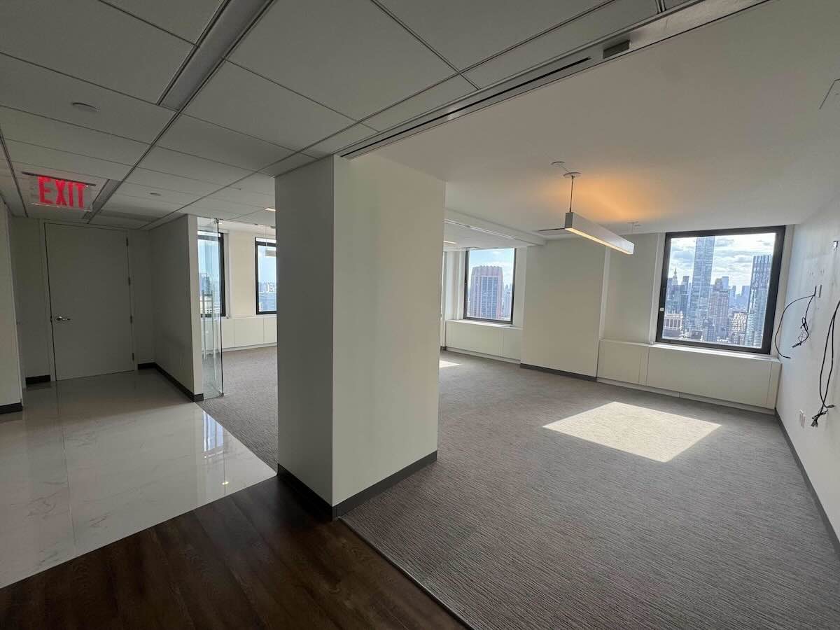 Modern office with city views, large windows, white walls, carpeted floor.