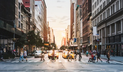 City street at sunset with people crossing, cars waiting, tall buildings.
