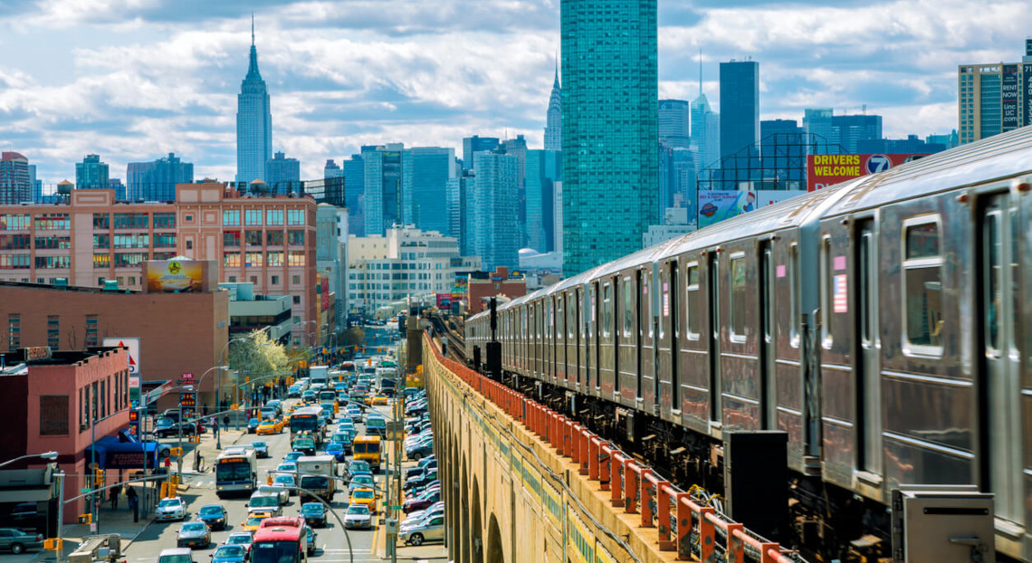Silver subway train on elevated tracks over busy city street, Empire State Building in the skyline.