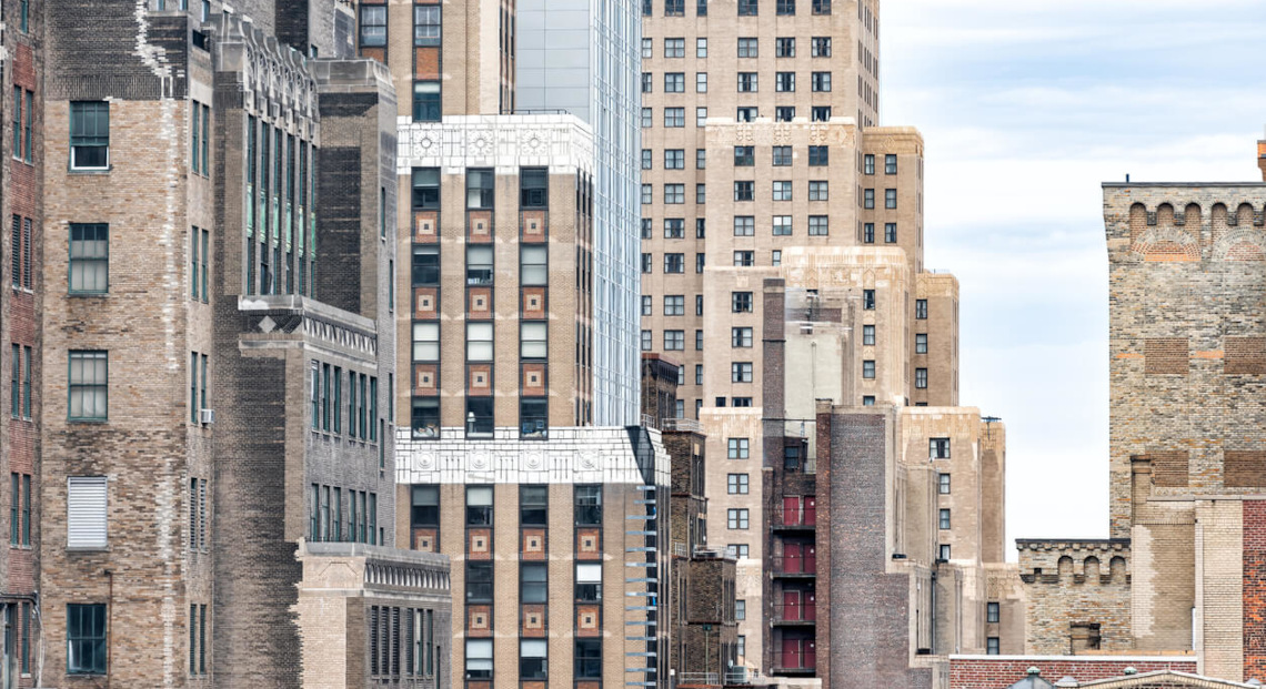 Cluster of tall brown city buildings with large windows under a bright, cloudy sky.