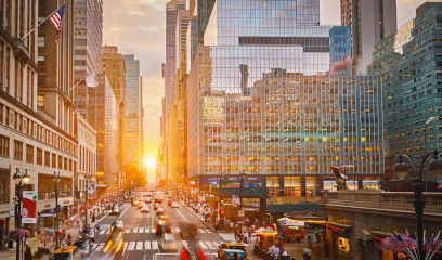 NYC street at sunset with skyscrapers, cars, people, and US flags glowing.