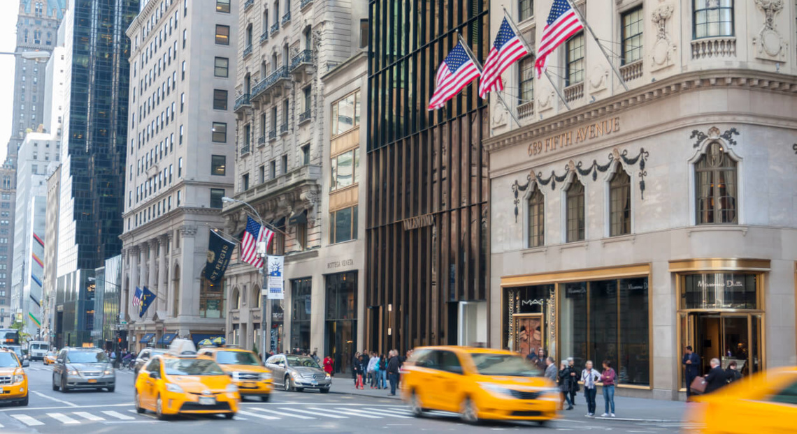 Yellow taxis on a busy NYC street lined with tall buildings; pedestrians walk nearby.