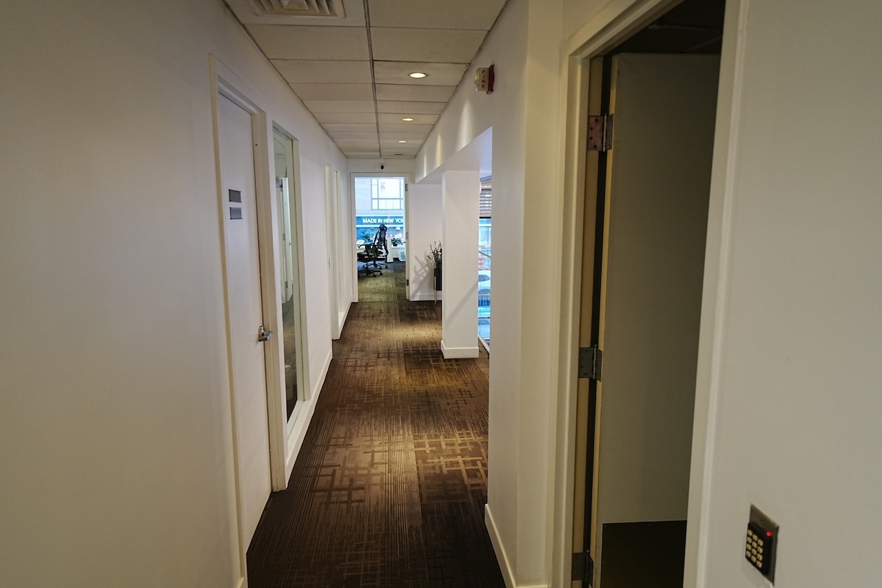 Office hallway with beige walls, brown carpet, doors, and workroom ahead.
