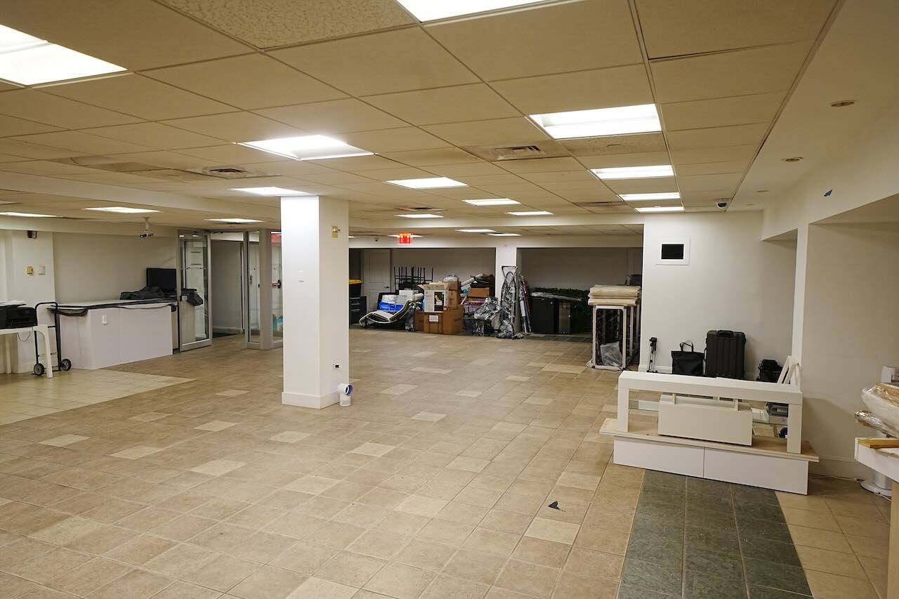 Empty tile-floored office with stacked furniture, white walls, glass door.