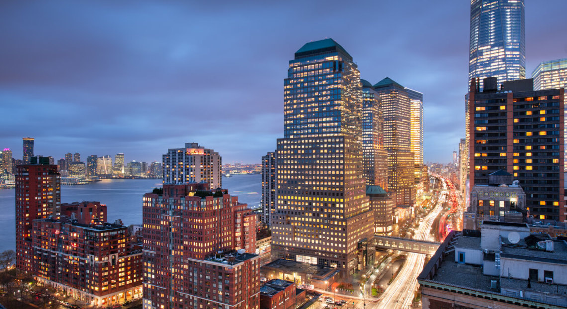 Dusk cityscape with illuminated skyscrapers, river reflections, and busy streets.