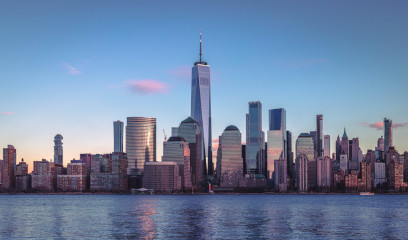 Sunset city skyline with modern skyscrapers, including One World Trade Center, reflected on calm water under a pink and blue sky.