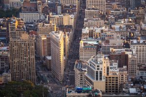 Flatiron Building in NYC, sunlight on its triangular shape, tall buildings and busy streets surround it.