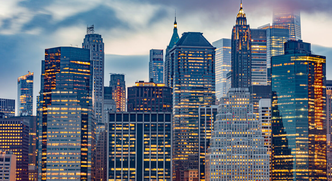 Modern skyscrapers with lit windows in NYC skyline at dusk under cloudy sky.