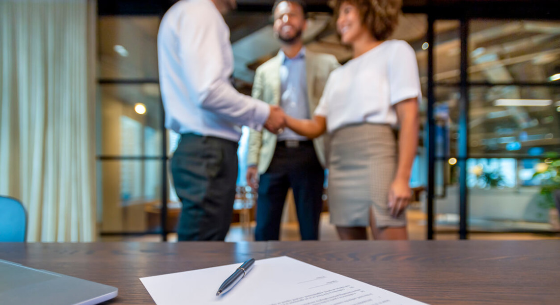 Pen and document on desk; three brokers shake hands in NYC office.
