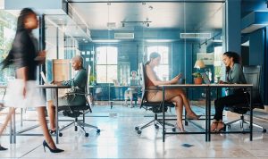 Modern office with glass walls, people at desks, plants, natural light.