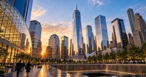 People walk near 9/11 Memorial pools, trees, and skyscrapers at sunset.