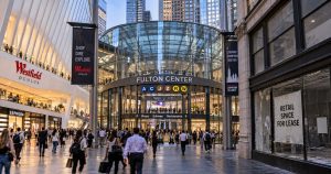 People walk by Fulton Center in NYC, with shops and subway signs nearby.