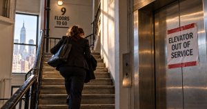 Person in business attire climbs stairs by “Elevator Out of Service” sign.